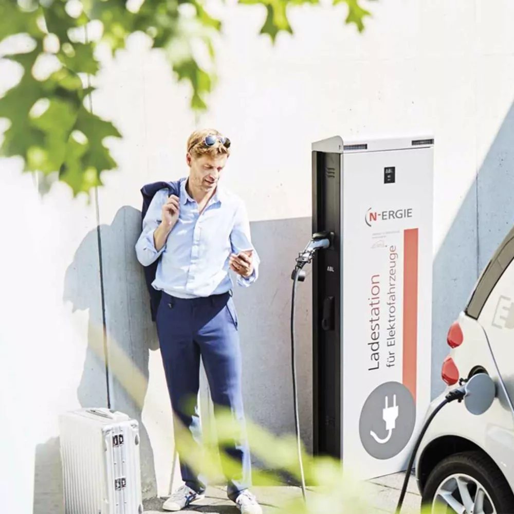 Man in business attire stands at an electric vehicle charging station, checking his smartphone while the car is charging.