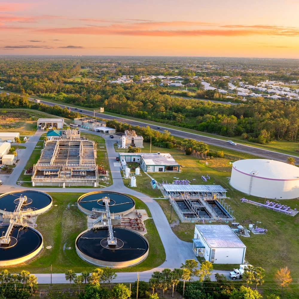 Aerial view of a water treatment facility with circular tanks, processing structures, roads, and surrounding greenery.